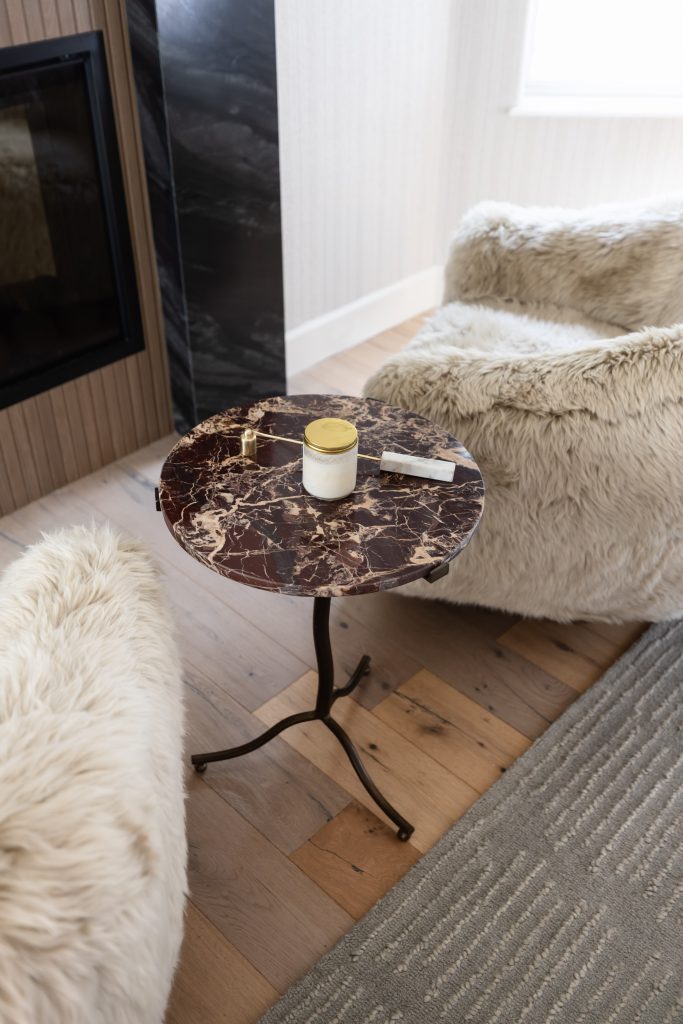 A small round brown marble side table with a candle, gold key, and white object sits between two beige faux fur chairs on wood flooring.— Living with Lolo.