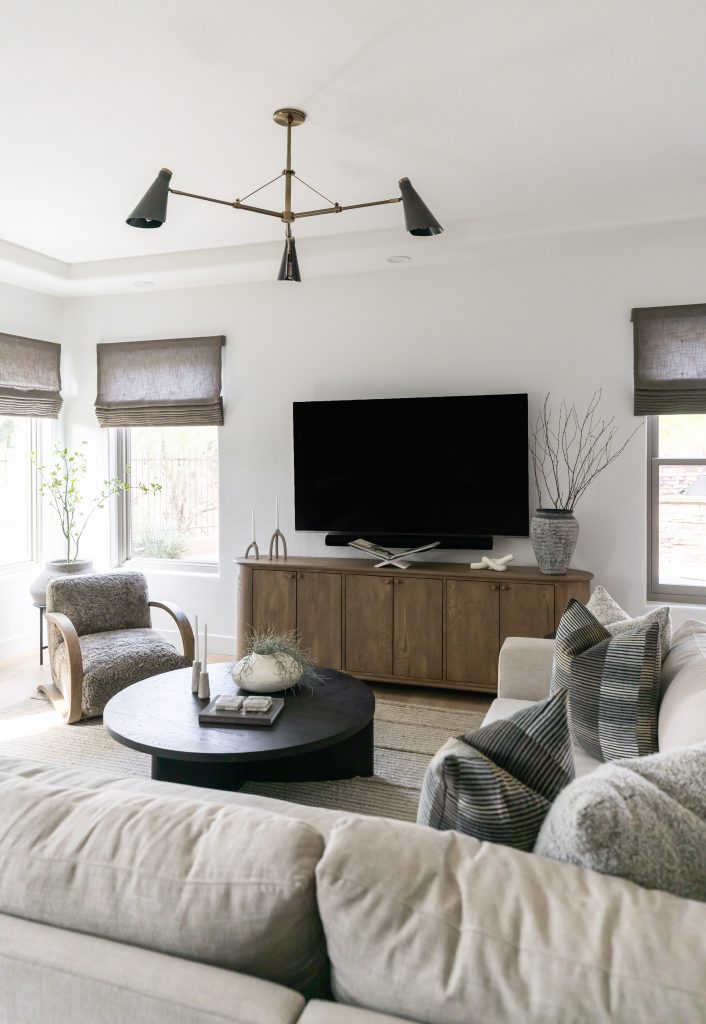 Modern living room featuring a beige sectional, black coffee table, wood media console, wall-mounted TV, armchair, and large windows with roman shades—Living with Lolo.