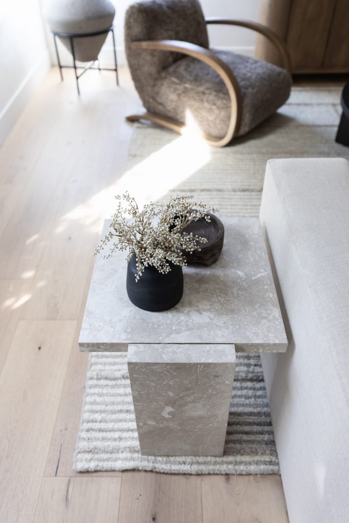 A marble side table with a black vase of dried flowers and a dark bowl is placed beside a beige sofa on a striped rug—Living with Lolo.
