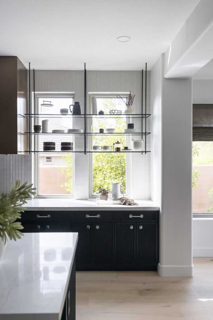 Modern kitchen with black lower cabinets, white countertop, open shelves on three windows, light wood flooring, and neutral walls—Living with Lolo.