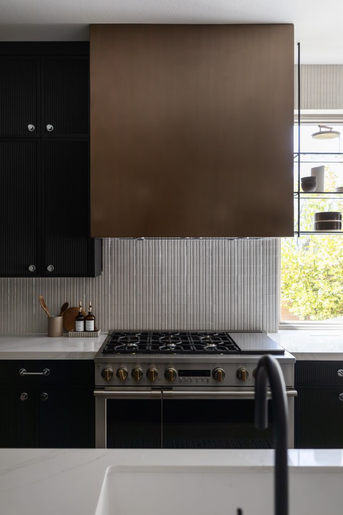 Modern kitchen with a brass range hood, black cabinets, stainless steel stove, white countertop, and window shelves on the right—Living with Lolo.