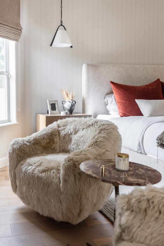 A beige chair, round marble side table with a candle, rust and gray pillows, and wooden nightstand sit under a hanging light—Living with Lolo.