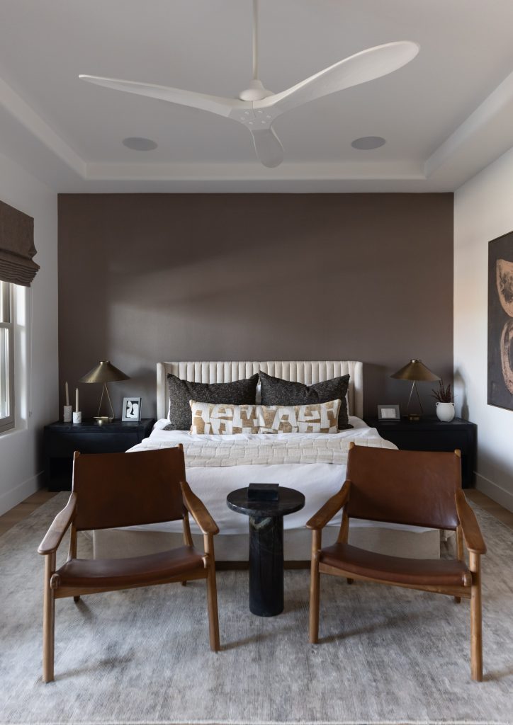 A modern bedroom with a large bed, two brown chairs, a round table, and a dark accent wall in neutral tones. — Living with Lolo.