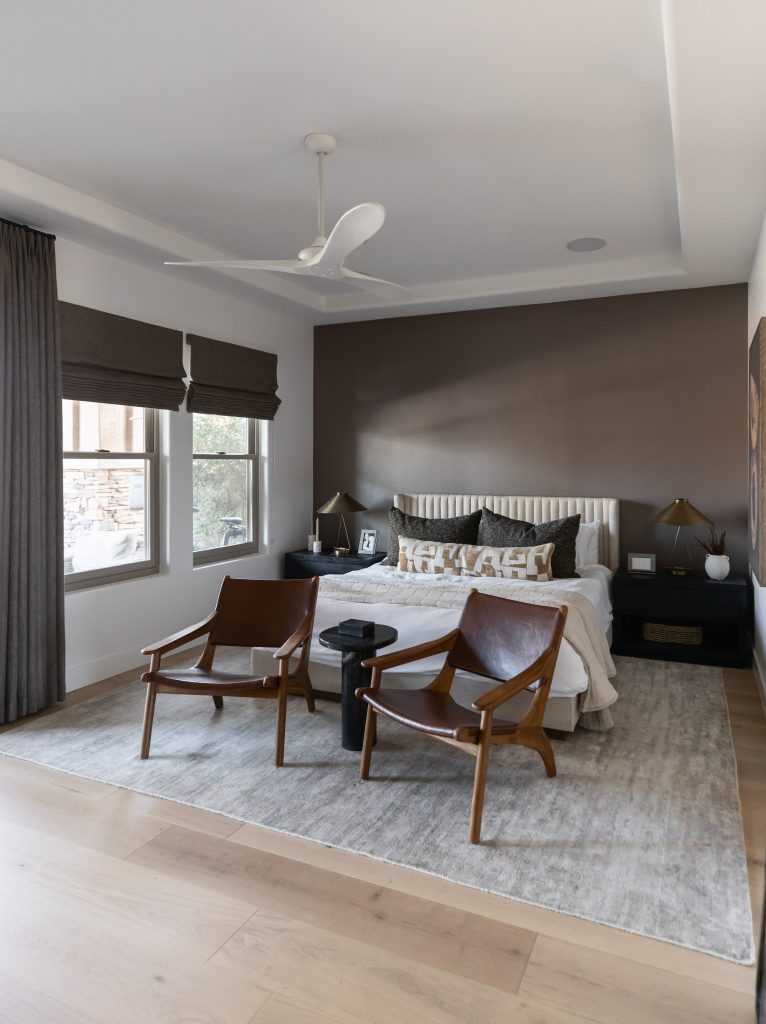Modern bedroom with a brown accent wall, white bedding, two brown leather chairs, a large rug, and windows with roman shades—Living with Lolo.