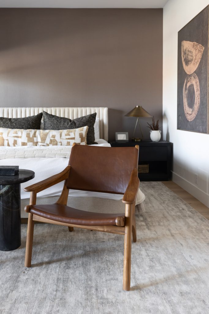 A modern bedroom with a leather accent chair, neutral bedding, a black nightstand, table lamp, and abstract wall art. — Living with Lolo.