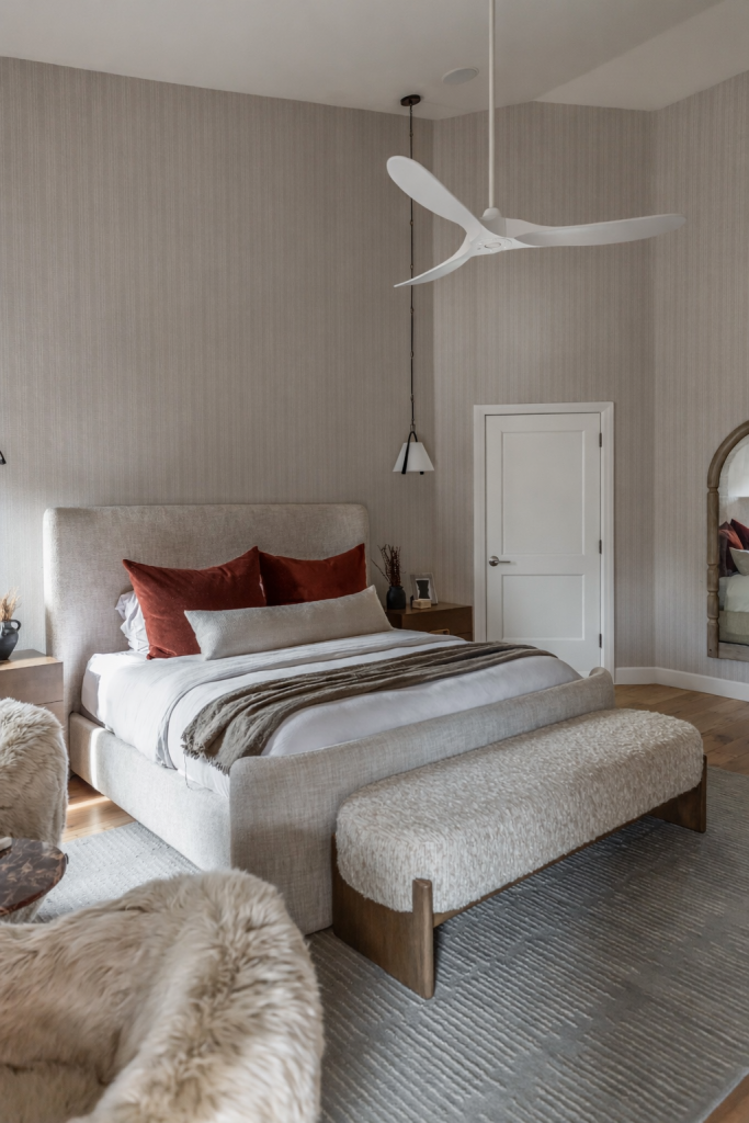 Modern bedroom with a neutral palette, large upholstered bed, red accent pillows, textured bench, fur throw, pendant lights, and ceiling fan. — Living with Lolo.