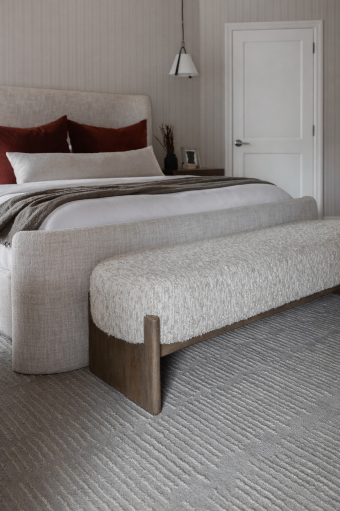 Modern bedroom with a beige upholstered bed, rust-colored pillows, a textured bench at the foot, and a closed white door—Living with Lolo.