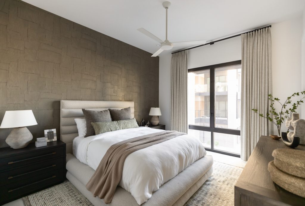 Modern bedroom with a textured accent wall, neutral upholstered bed, two bedside tables, large curtained window, and ceiling fan—Living with Lolo.