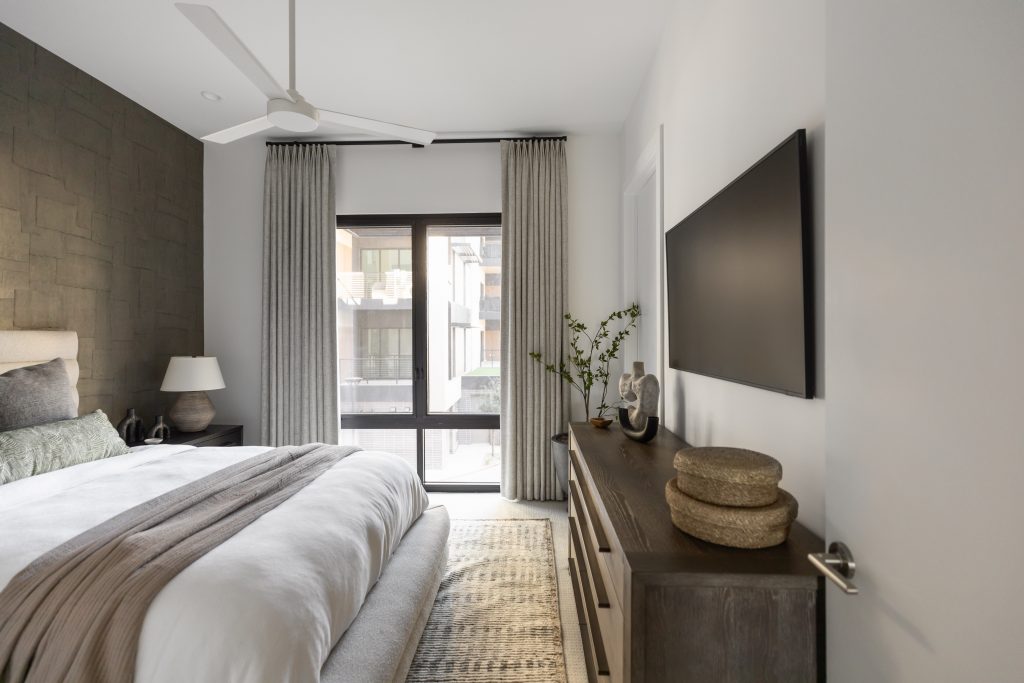 Modern bedroom featuring a large bed with gray bedding, a dresser, wall-mounted TV, lamp, potted plant, and curtained window—Living with Lolo.