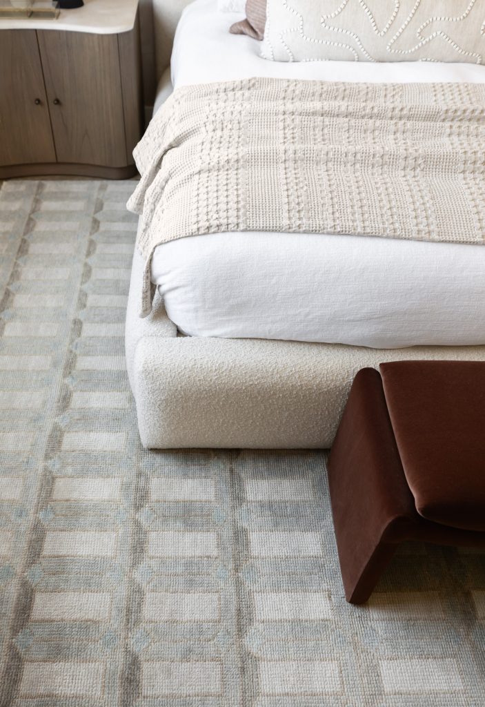 A beige textured blanket is folded on a white upholstered bed beside a patterned rug and a brown bench.— Living with Lolo.