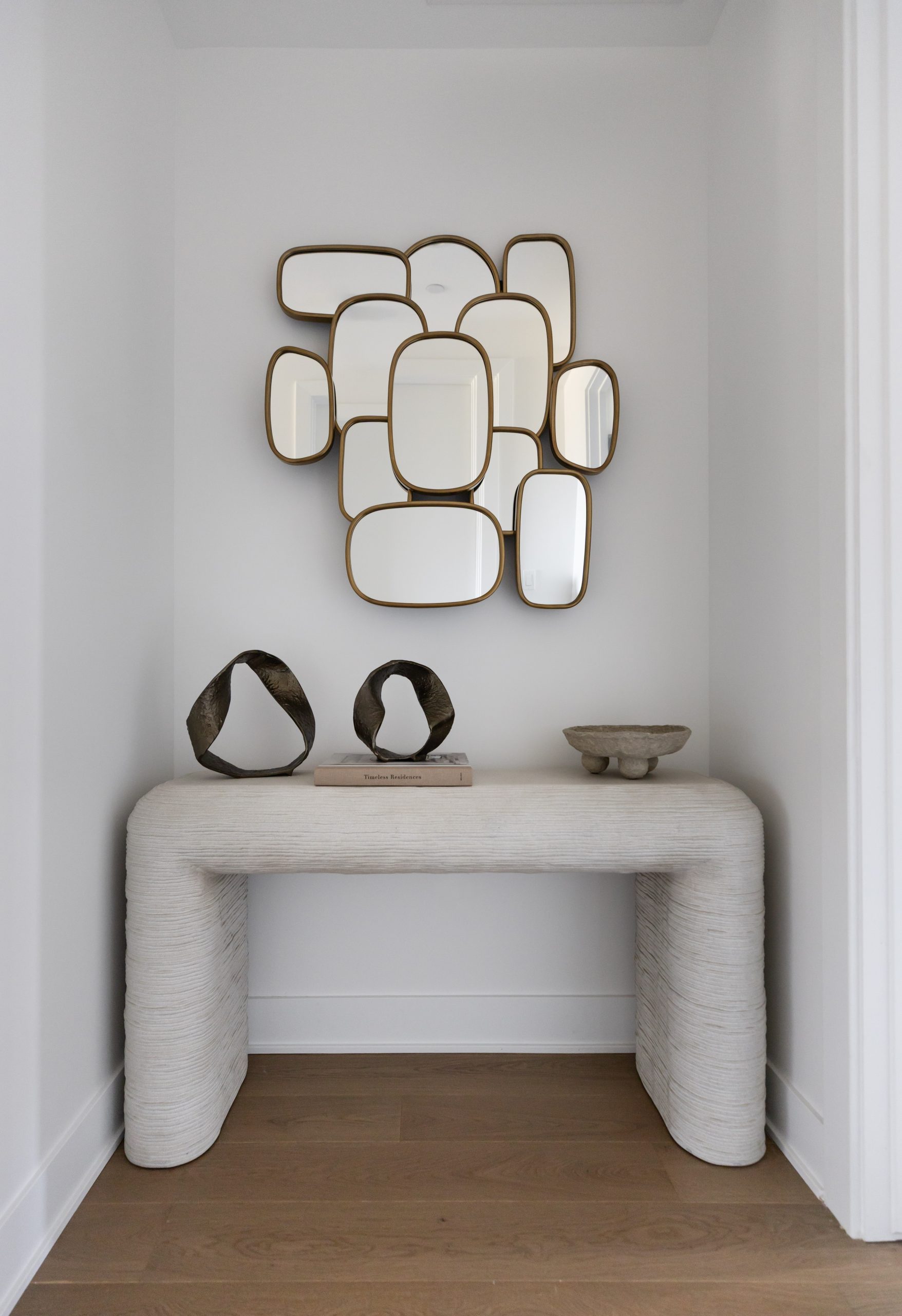 A modern console table with sculptural decor sits below a multi-panel mirror in a white alcove with wooden flooring—Living with Lolo.
