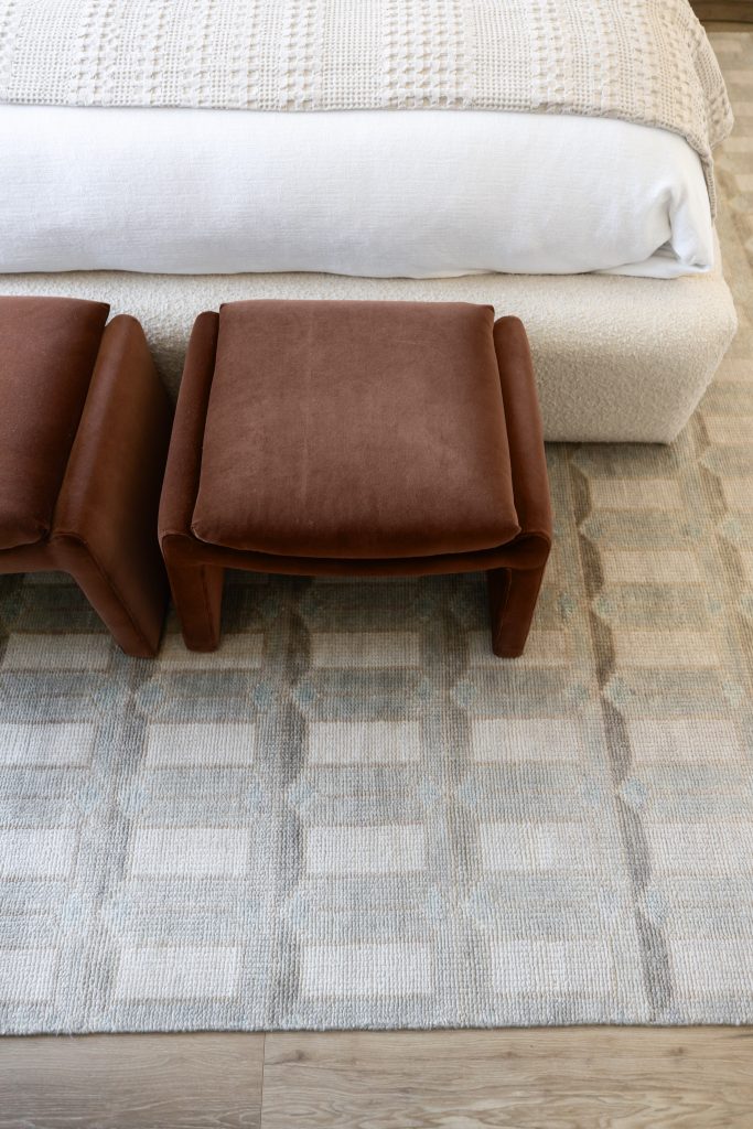 Two brown upholstered ottomans sit side by side at the foot of a bed with a textured beige blanket on a patterned area rug—Living with Lolo.