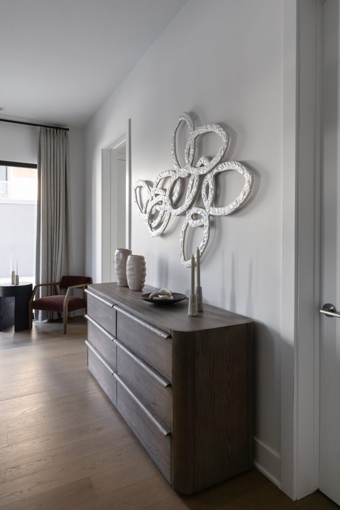 A hallway with a dark wood dresser, vases, two candlesticks, an abstract silver wall sculpture, light wood floors, and neutral drapes — Living with Lolo.