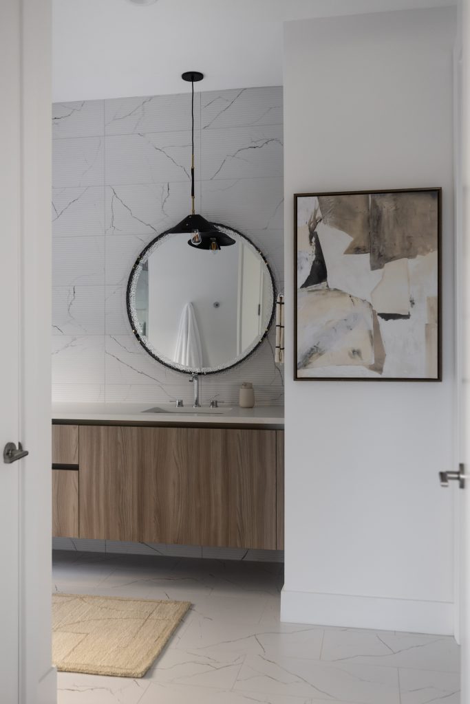 Modern bathroom with a round mirror above a wood vanity, marble-patterned walls and floor, black light fixture, and abstract wall art — Living with Lolo.