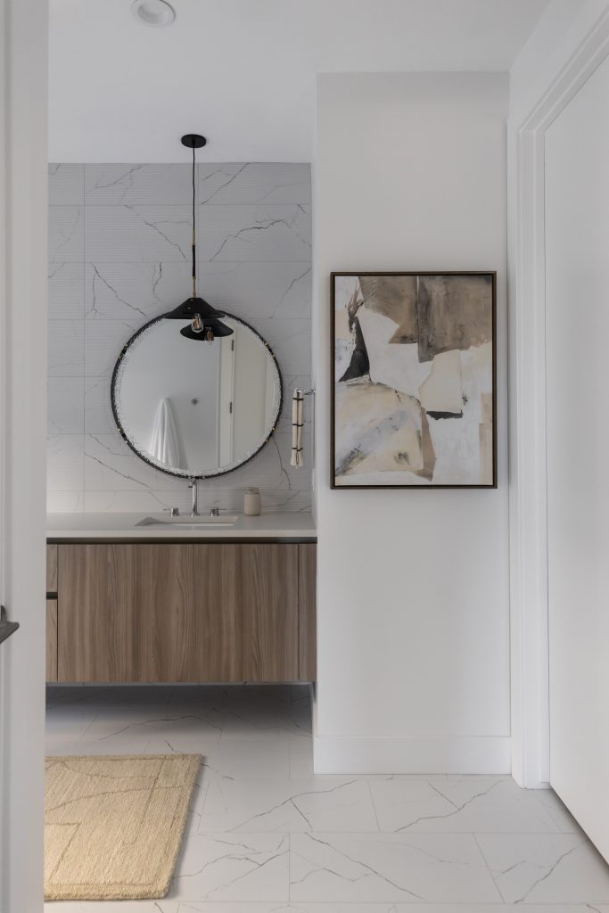 Modern bathroom with round mirror over wood vanity, abstract wall art, marble tile walls and floor, and a beige rug—Living with Lolo.