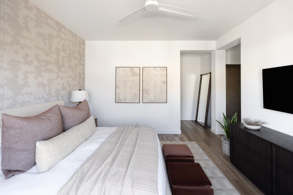 A modern bedroom with a large bed, two brown benches, a dresser, wall-mounted TV, potted plant, floor mirror, and abstract art.— Living with Lolo.