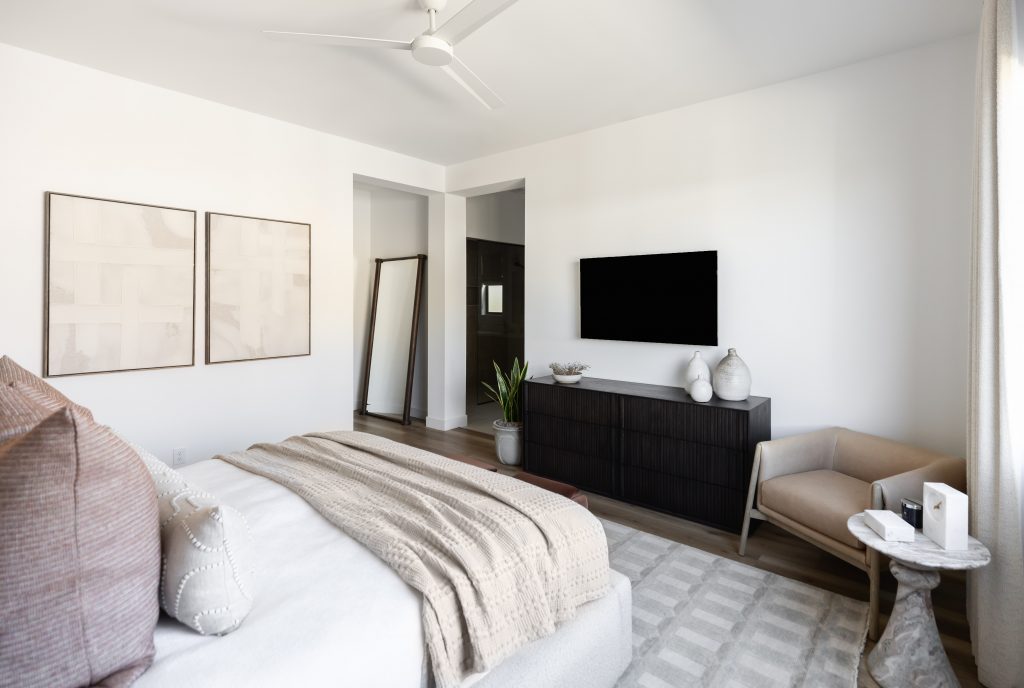 Minimalist bedroom with a bed in neutral bedding, wall art, dresser, wall-mounted TV, lounge chair, side table, plant, and mirror.— Living with Lolo.