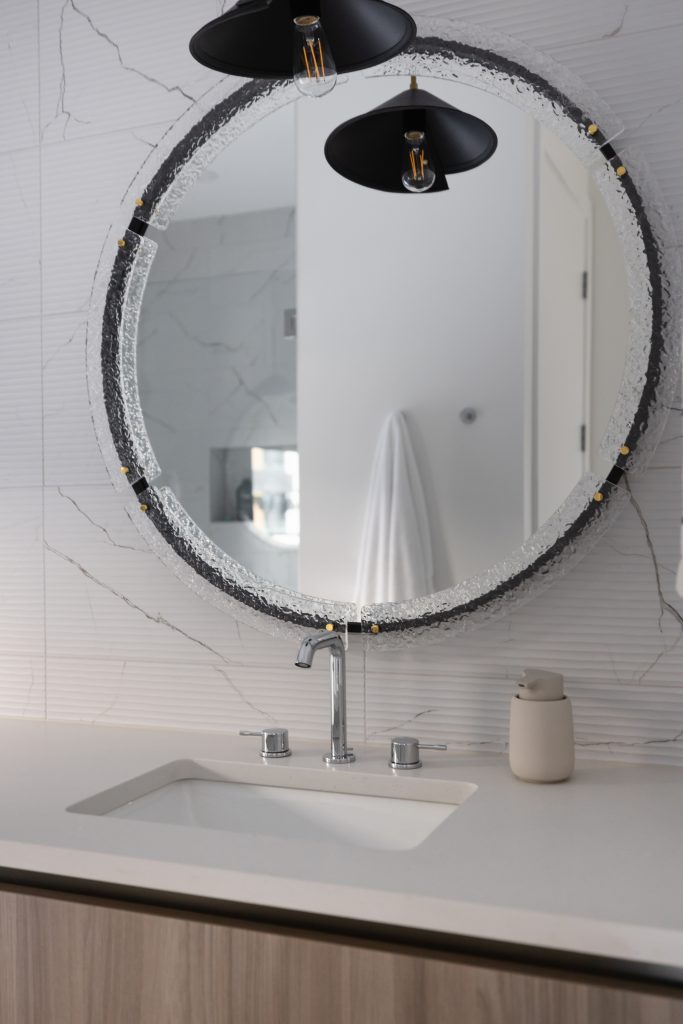 Modern bathroom sink with a round textured mirror above, a soap dispenser on the countertop, and a towel reflected in the mirror—Living with Lolo.