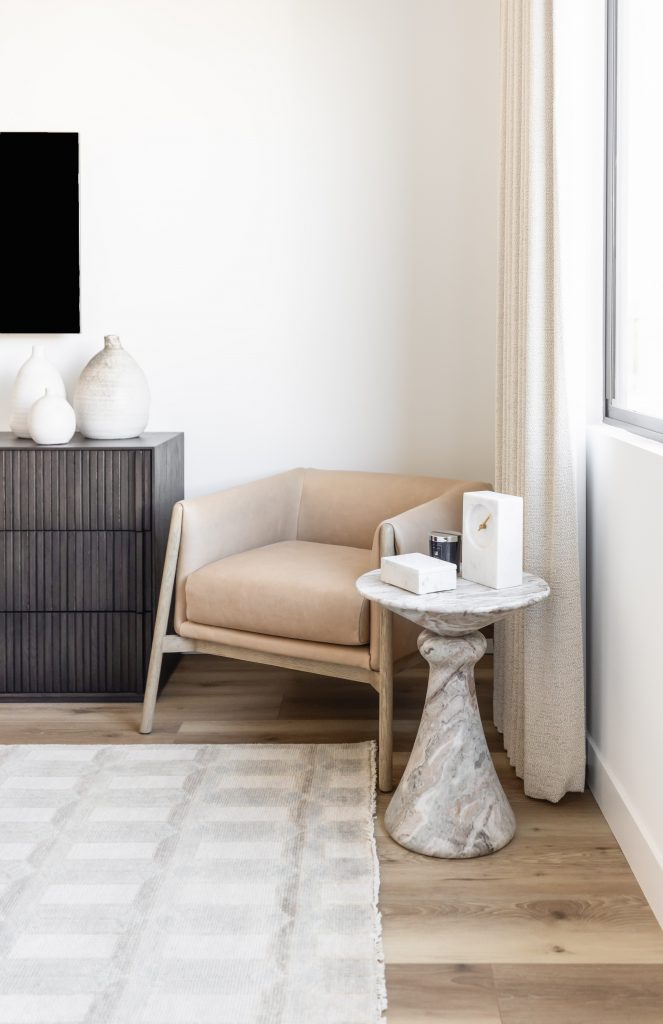 A tan armchair, marble side table, dark wood dresser, vases, and patterned rug sit on wood floors beside a window. — Living with Lolo.