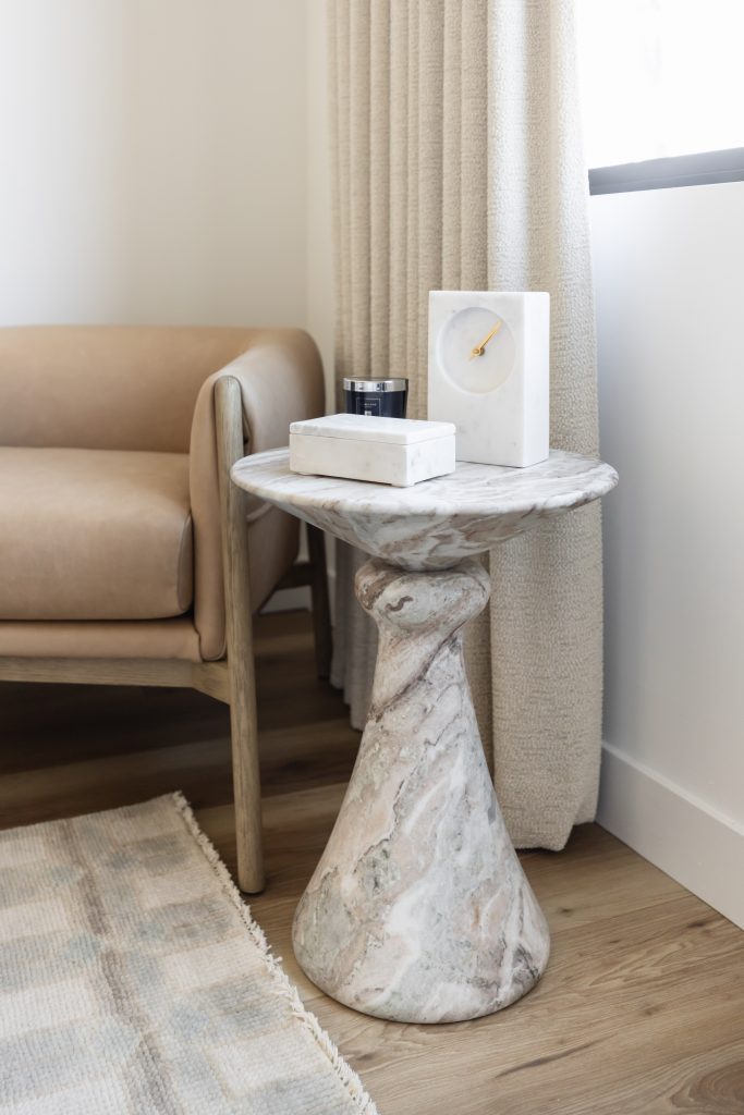 A marble side table holding a clock, candle, and rectangular box stands beside a tan armchair in a neutral-toned room—Living with Lolo.
