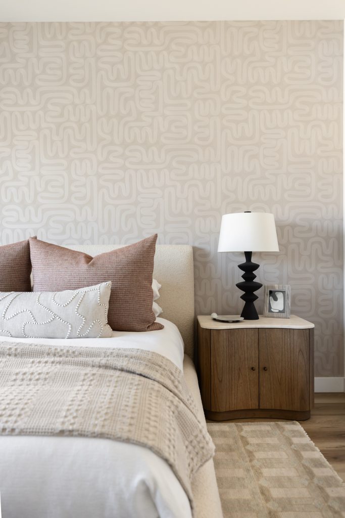 A modern bedroom corner with a beige patterned wall, bed, textured bedding, wooden nightstand, black lamp, and framed photo—Living with Lolo.