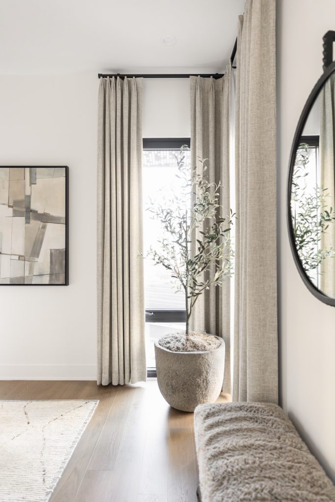 A potted plant sits next to tall beige curtains in a room with light wood floors, a bench, and an abstract painting— Living with Lolo.