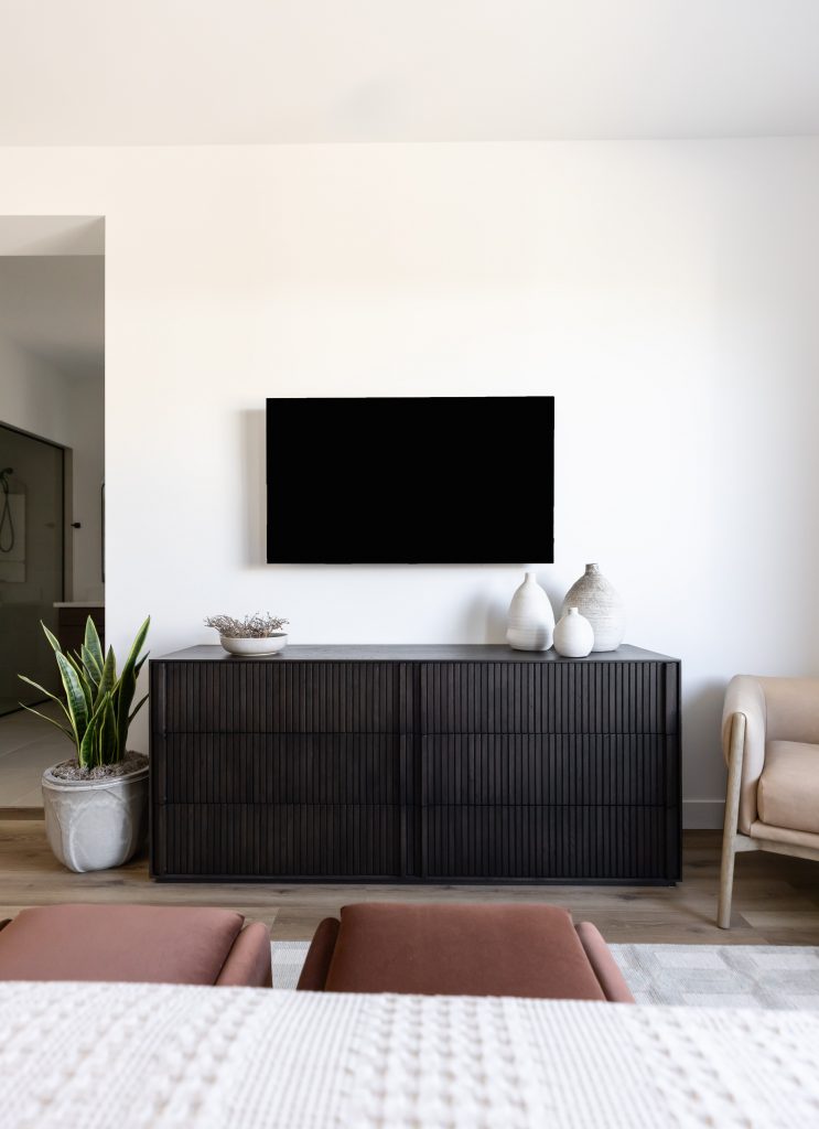 A wall-mounted flat-screen TV hangs above a dark sideboard with vases and a bowl. A potted plant is beside a light chair — Living with Lolo.