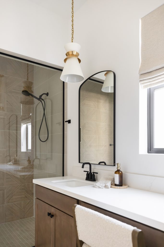 Modern bathroom featuring a glass shower, single sink vanity with black fixtures, wall mirror, pendant light, and window shade—Living with Lolo.