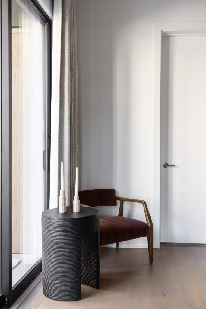 A small round black side table with two candles sits beside a brown armchair, near a window with a white door and gray curtain—Living with Lolo.