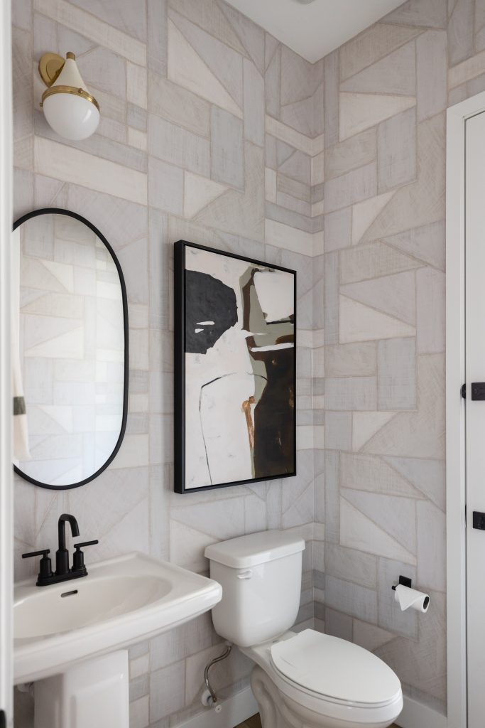 A bathroom with a white sink, black faucet, oval mirror, abstract wall art, toilet, and geometric-patterned wallpaper. — Living with Lolo.