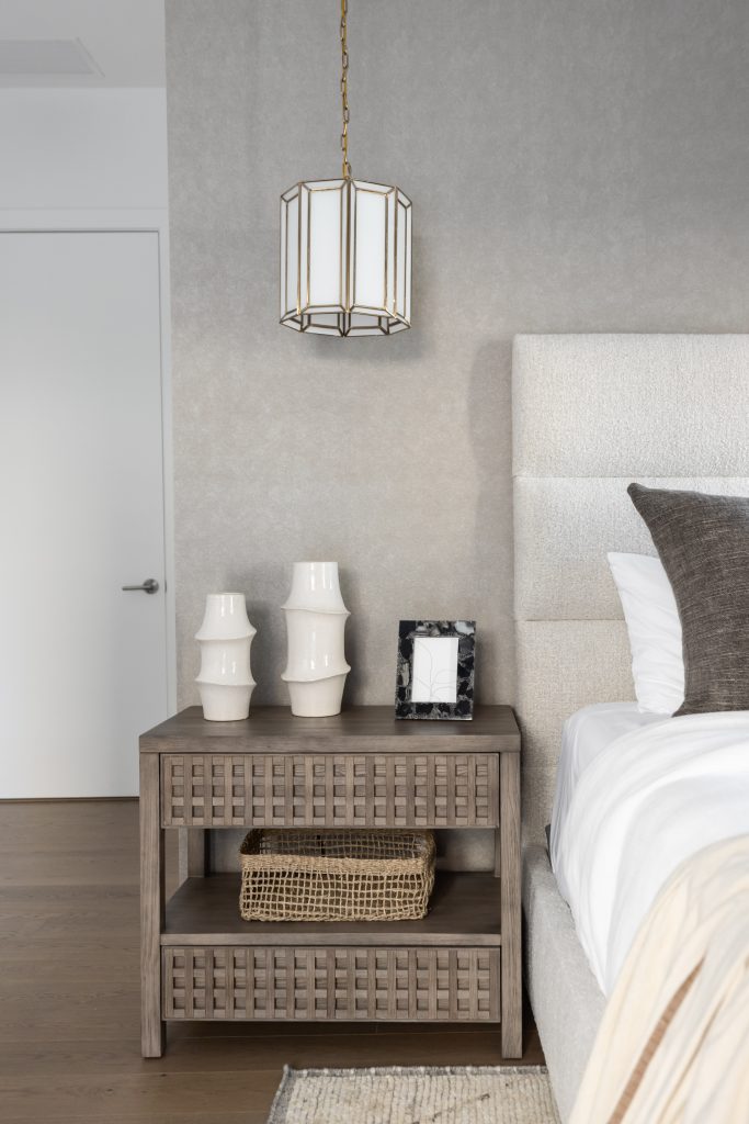 A wooden nightstand with two white vases, a black picture frame, and a woven basket sits beside a bed with gray and white bedding; a pendant light hangs above—Living with Lolo.