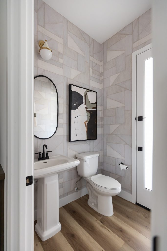 Modern bathroom with geometric wallpaper, pedestal sink, oval mirror, toilet, abstract wall art, wood floor, and wall sconce—Living with Lolo.