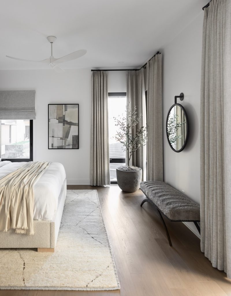 A modern bedroom features light walls, sheer curtains, a bed with a beige throw, abstract wall art, a round mirror, and light wood floors—Living with Lolo.