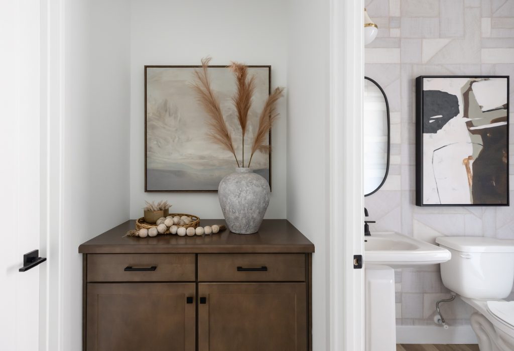 A brown cabinet with a gray vase of dried pampas grass, decorative beads, and a landscape painting sits in a nook beside a bathroom — Living with Lolo.