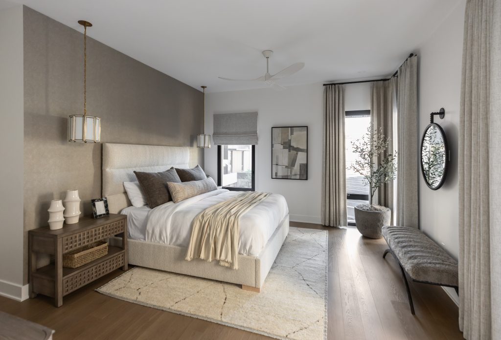 Modern bedroom featuring a large bed with neutral bedding, pendant lights, wood nightstand, bench, abstract art, and large curtained windows—Living with Lolo.