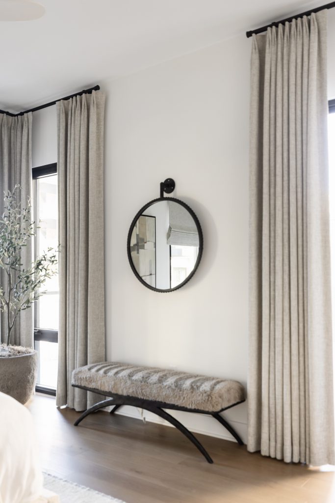 A round mirror is mounted above an upholstered bench with metal legs, set between two tall windows with light curtains—Living with Lolo.