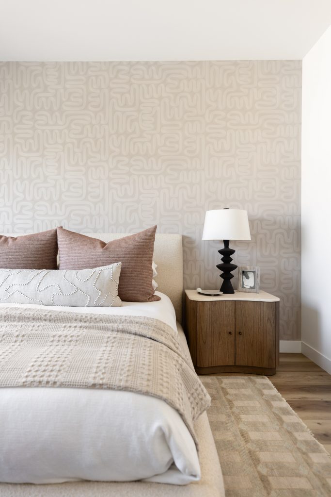 A neatly made bed with neutral bedding sits next to a wooden nightstand with a black lamp and photo frame against a cream accent wall—Living with Lolo.