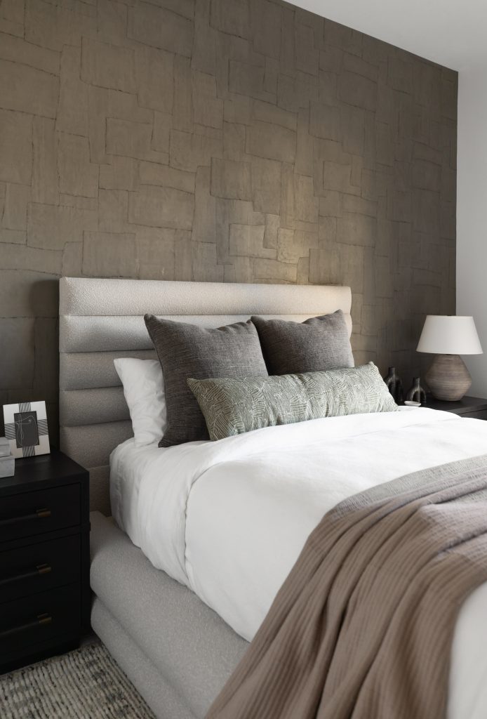 A bedroom with a textured gray accent wall, upholstered bed, white bedding, gray and green pillows, beige blanket, black nightstands, and a table lamp—Living with Lolo.