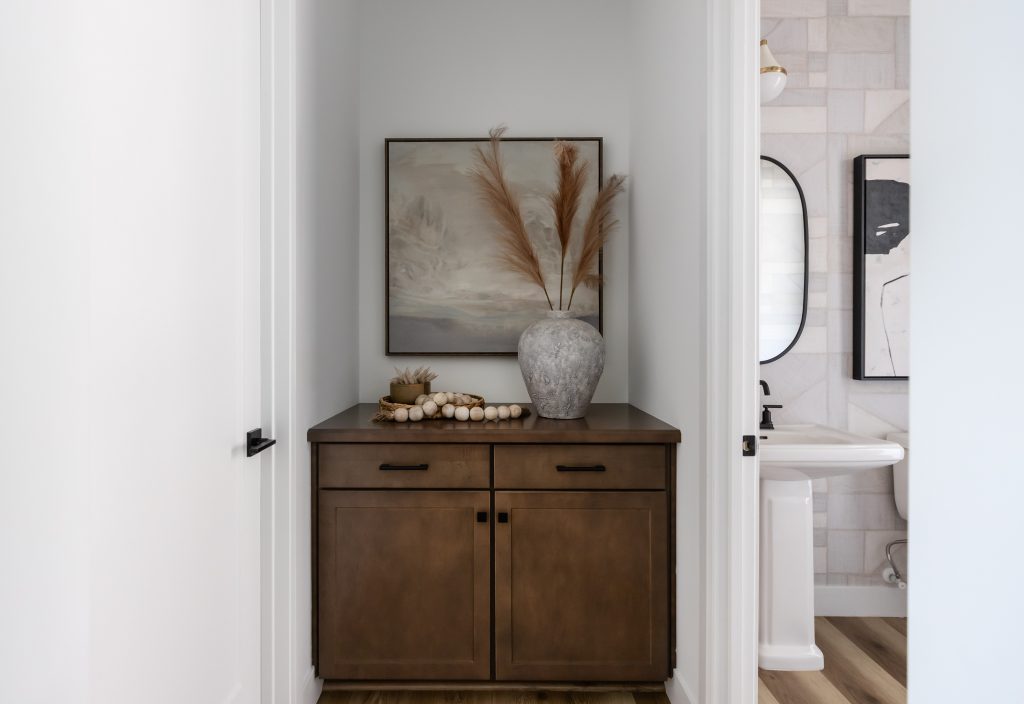 A wooden cabinet with a vase of dried grasses and a tray of beads stands below a landscape painting in a hallway near a bathroom. — Living with Lolo.