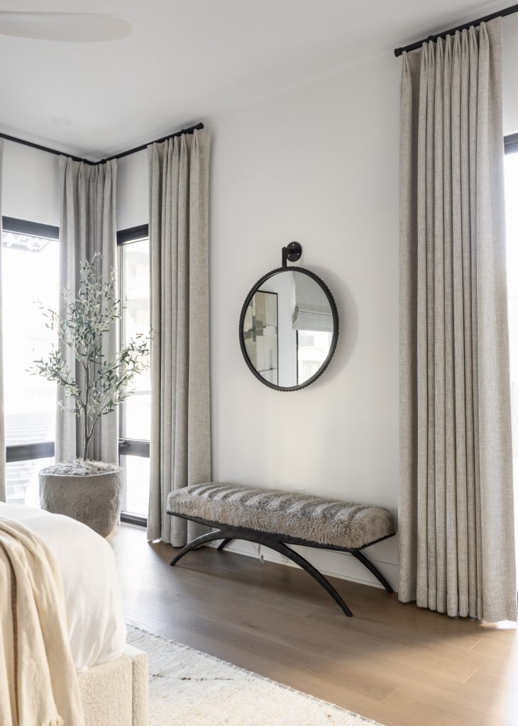 A bedroom corner features tall beige curtains, a round wall mirror, a textured bench, a large potted plant, and light wood flooring. — Living with Lolo.