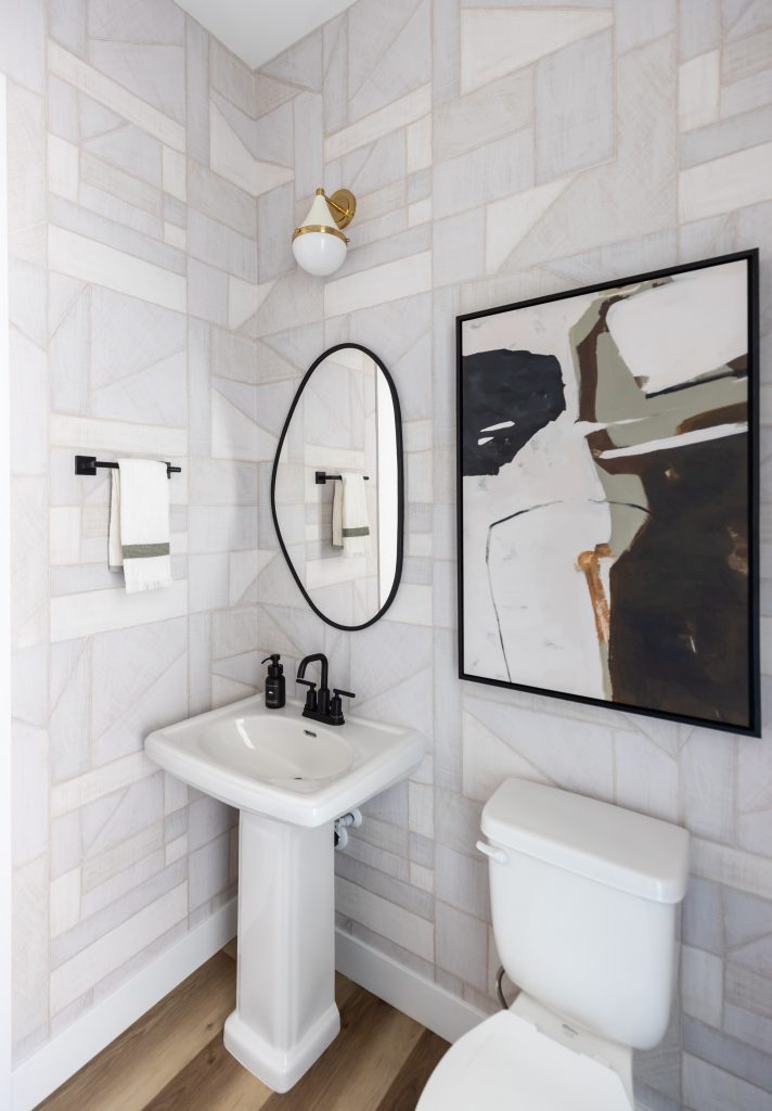 A small bathroom with a white pedestal sink, black fixtures, oval mirror, abstract wall art, towel rack, and patterned wallpaper — Living with Lolo.