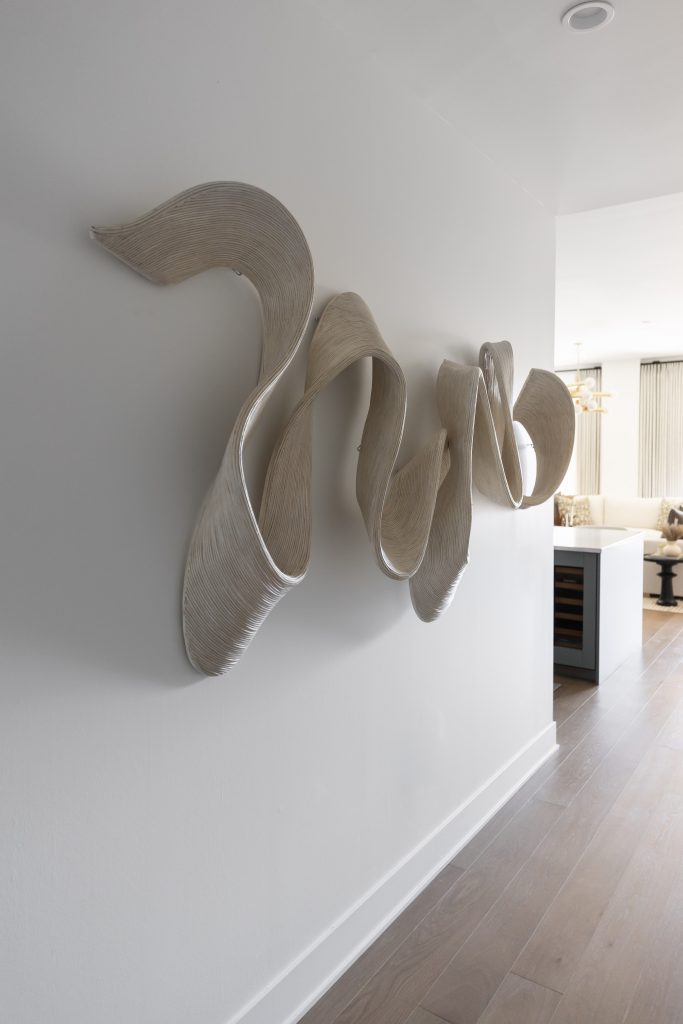 A wall-mounted abstract sculpture with smooth, ribbon-like curves is displayed in a modern hallway—Living with Lolo.