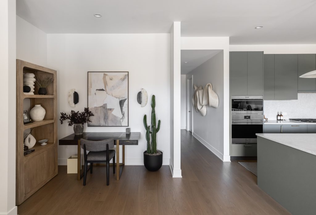 Modern interior featuring a small desk and chair, abstract wall art, tall potted cactus, built-in shelves, and a minimalist kitchen—Living with Lolo.