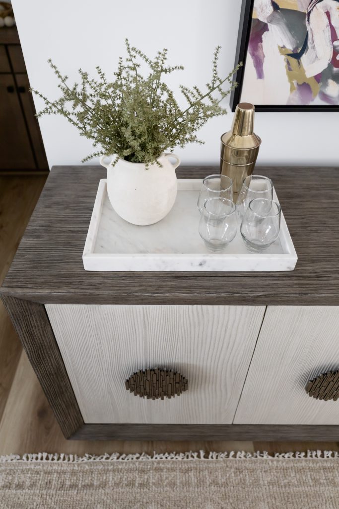 A white vase with greenery, a gold cocktail shaker, and three empty glasses rest on a marble tray on a wooden cabinet—Living with Lolo.