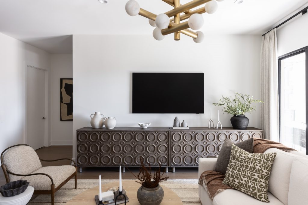 Modern living room featuring a patterned sideboard, wall-mounted TV, vases, beige armchair, light sofa with pillows, and gold chandelier—Living with Lolo.