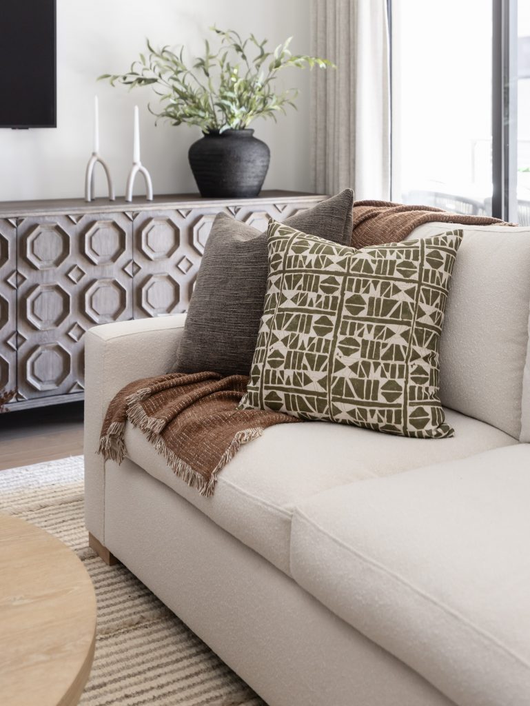 A white sofa with a patterned green pillow, brown throw, neutral rug, and a geometric wooden sideboard with a black vase of greenery. — Living with Lolo.