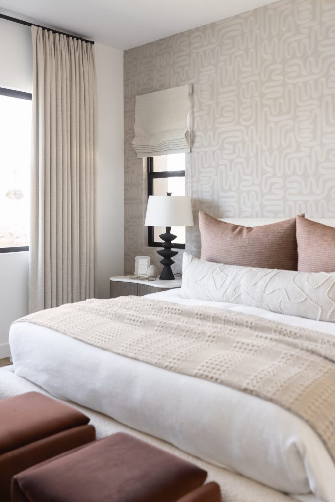 Neutral-toned bedroom with patterned wallpaper, a bed with light bedding and brown pillows, bedside table, and two brown ottomans—Living with Lolo.