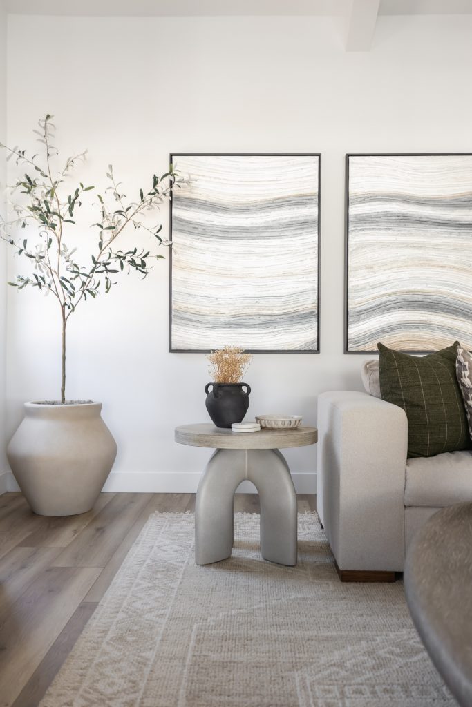 A beige sofa, round side table, large potted plant, and two abstract wave art pieces against a white wall. — Living with Lolo.