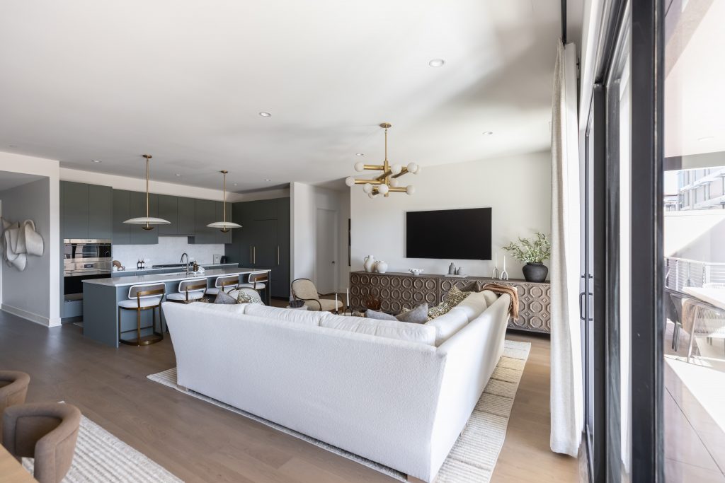 Open-plan living room and kitchen with a white sofa, wall-mounted TV, kitchen island with barstools, pendant lights, and large windows. — Living with Lolo.