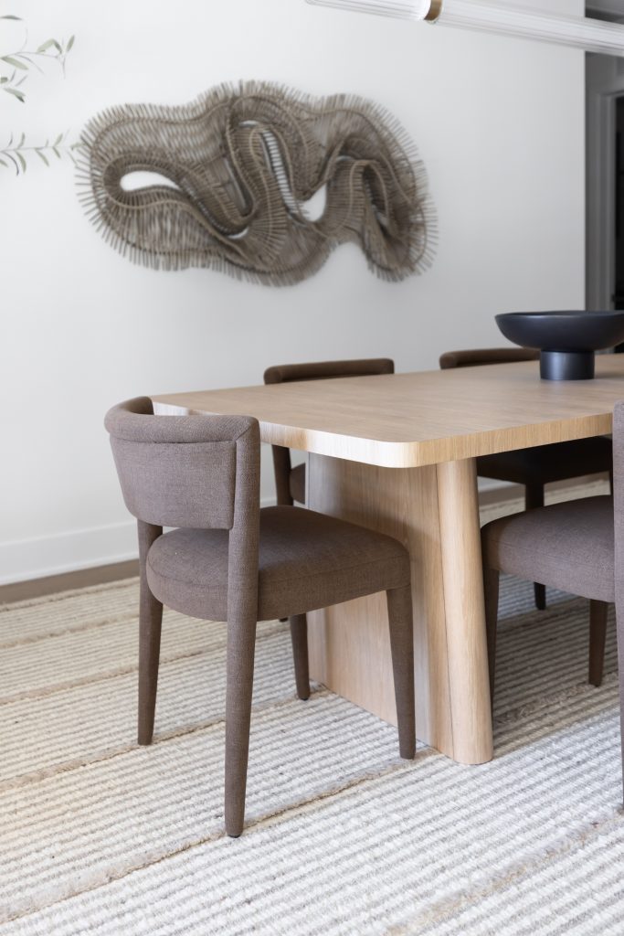 A light wooden dining table with four brown upholstered chairs and a black bowl sits beneath abstract wall art.— Living with Lolo.